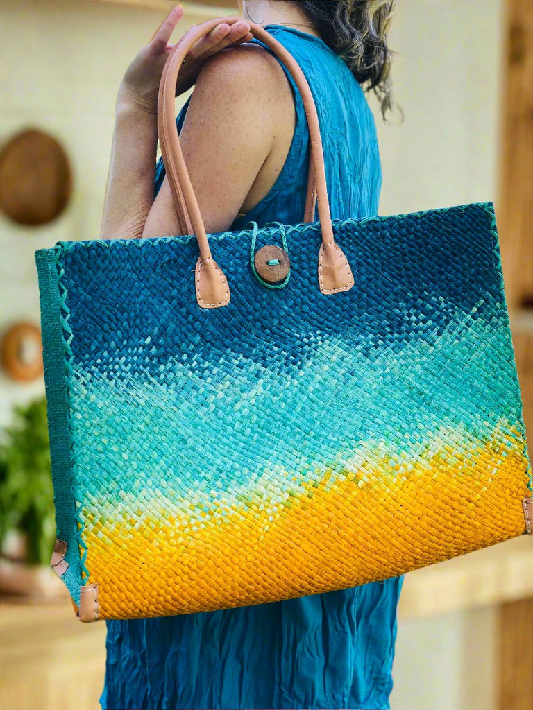 Model wearing Holden Turquoise Ombre Zafran Large Straw Beach Bags handmade dip dyed raffia woven into a three part horizontal ombre pattern of turquoise blue on the top, seafoam blue/green in the middle and both sides, and saffron yellow on the bottom with cross stitch edging, wooden button, and leather handles - Shebobo