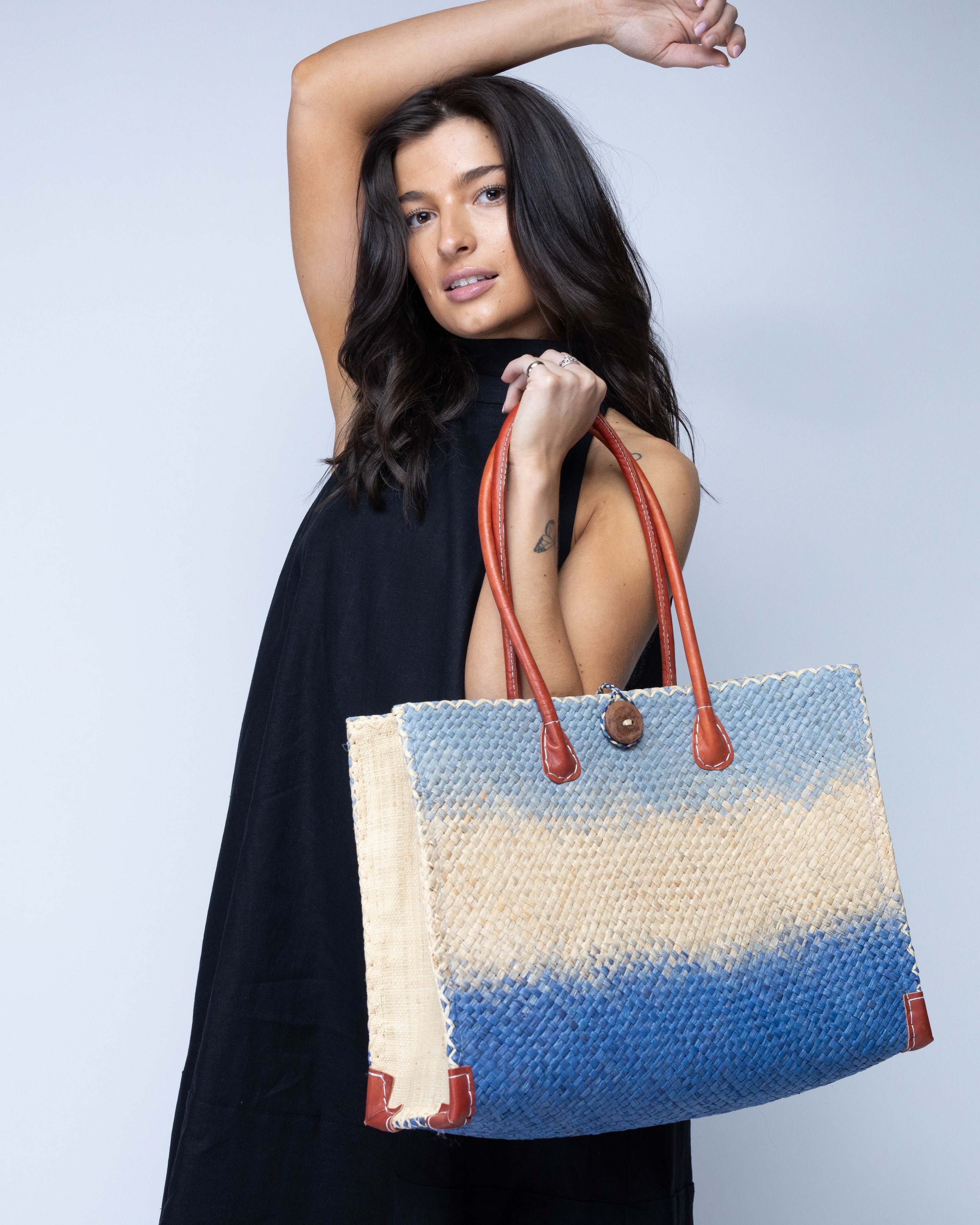 Model wearing Small Holden Denim Ombre Zafran Straw Beach Bags handmade dip dyed raffia woven into a three part horizontal ombre pattern of denim retro/light/blue, natural straw color, and dusk dark/navy/blue with cross stitch edging, wooden button, and leather handles - Shebobo