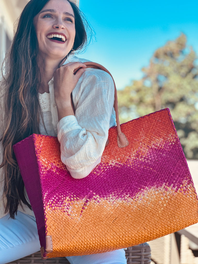 Model wearing Holden Fuchsia Ombre Zafran Straw Beach Bags handmade dip dyed raffia woven into a three part horizontal ombre pattern of coral orange/red on the top, fuchsia pink in the middle and both sides, and saffron yellow on the bottom with cross stitch edging, wooden button, and leather handles - Shebobo