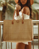 Model wearing Zafran Large Straw Beach Bag with Plastic Liner handmade loomed raffia in natural straw color with contrasting cross stitch edge binding, wood button closure, and leather handles & feet plus assorted print plastic lining - Shebobo