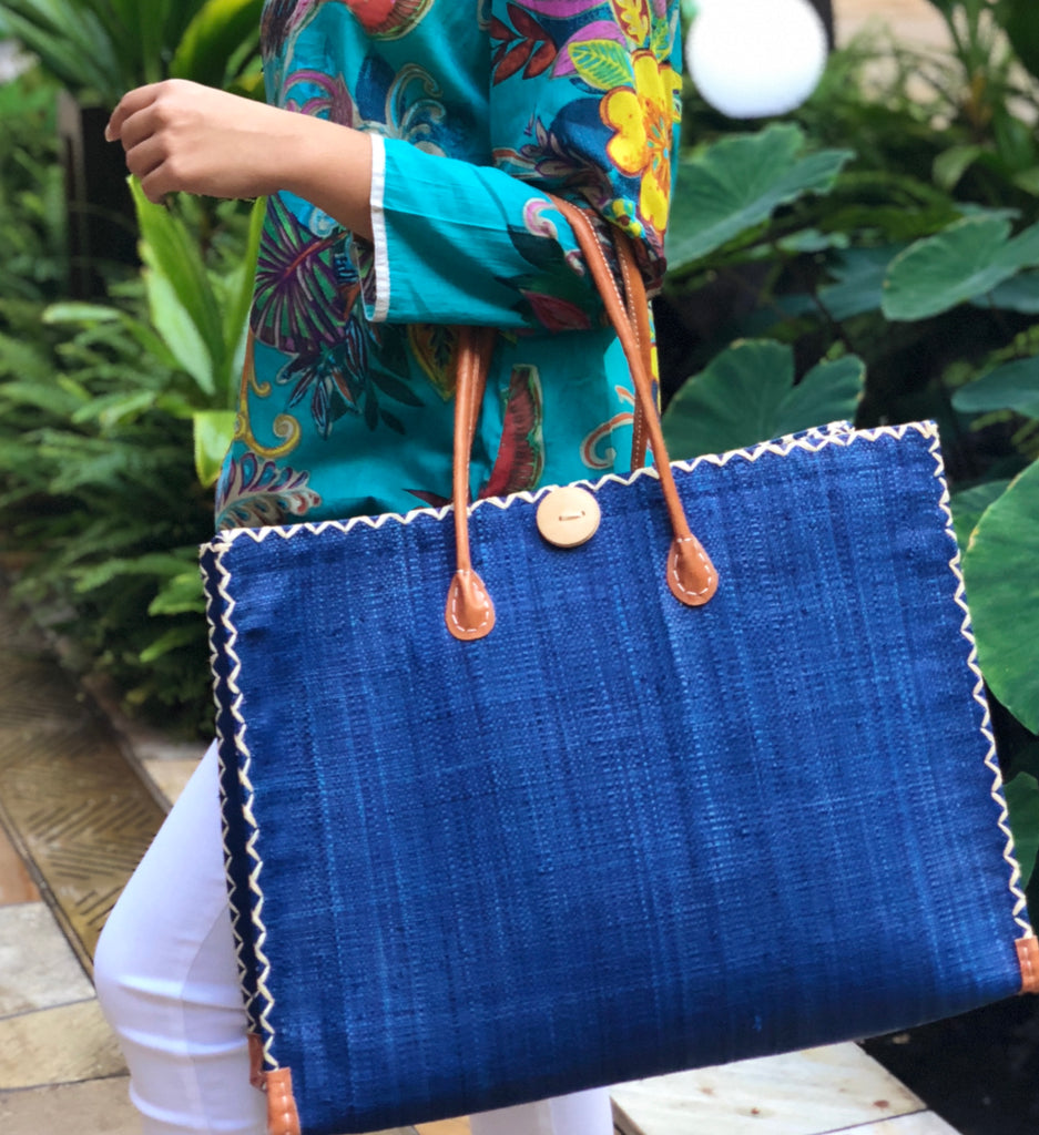 Model wearing Zafran Large Straw Beach Bag with Plastic Liner handmade loomed raffia in Navy deep blue with contrasting cross stitch edge binding, wood button closure, and leather handles & feet plus assorted print plastic lining - Shebobo