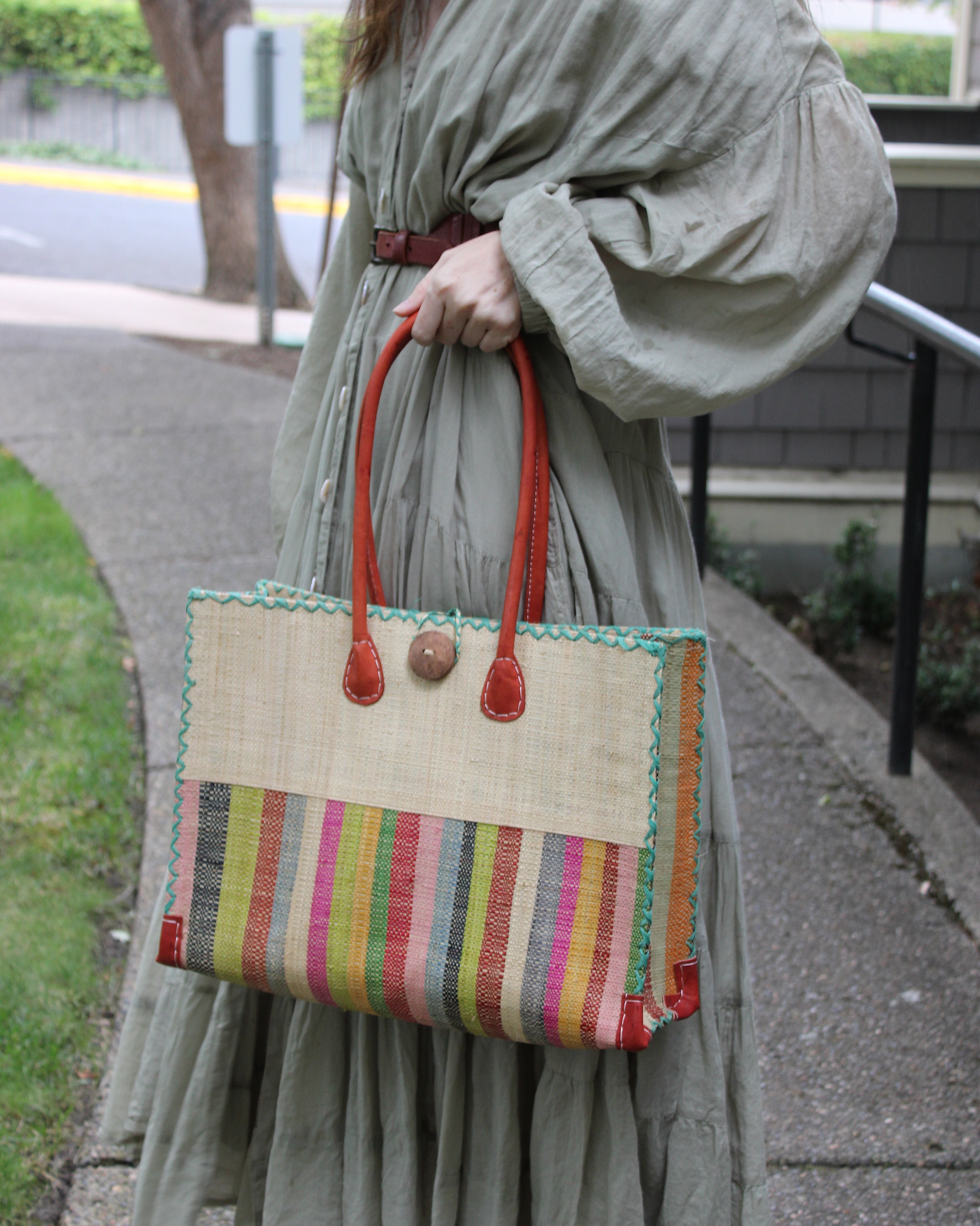 Model wearing Zafran Two Tone Wide Stripe Lollipop Stripe Straw Beach Bag handmade loomed raffia palm fiber in a solid hue of Natural straw color on the top half of the bag and a multicolor multi-width vertical stripe pattern of black, lime green, grass green, red, light/soft/baby pink, yellow, grey, whisky caramel/red/brown, light blue, fuchsia hot/bright/barbie/pink, and natural straw color on the bottom half of the bag with leather handles and feet and matching crosshatch stitch edging shoulder bag - She