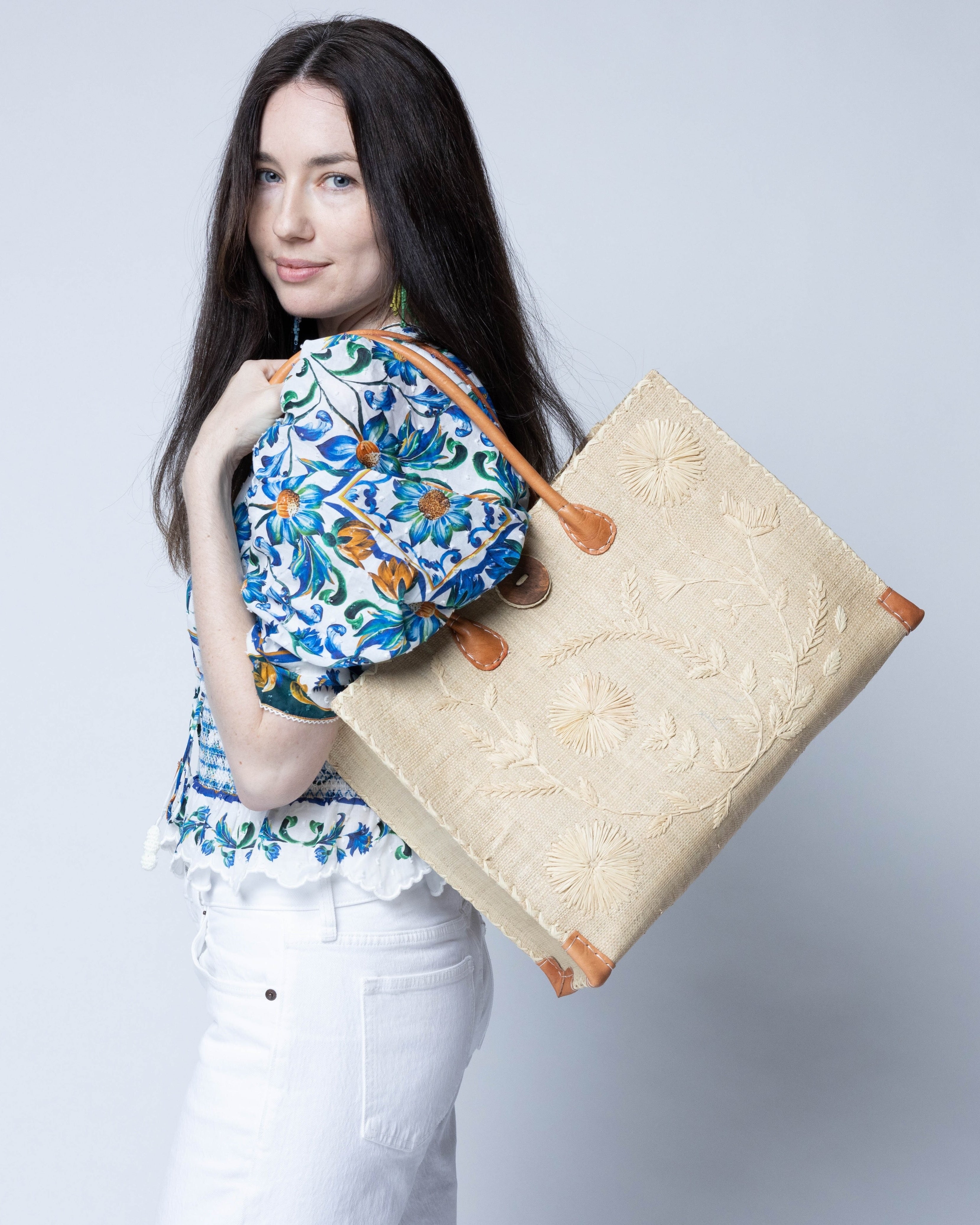 Model wearing Zafran Natural Embroidered Straw Beach Bag with Floral Pattern handmade natural raffia palm fiber in a solid hue of natural straw color with asymmetrical floral leaf, vine, and flower pattern in Natural with matching cross-stitch edging, leather handles, and wooden button closure shoulder bag handbag tote - Shebobo