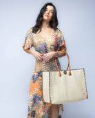Model wearing Large Zafran Straw Beach Bag with Plastic Liner handmade loomed raffia in natural straw color with contrasting cross stitch edge binding, wood button closure, and leather handles & feet plus assorted print plastic lining - Shebobo