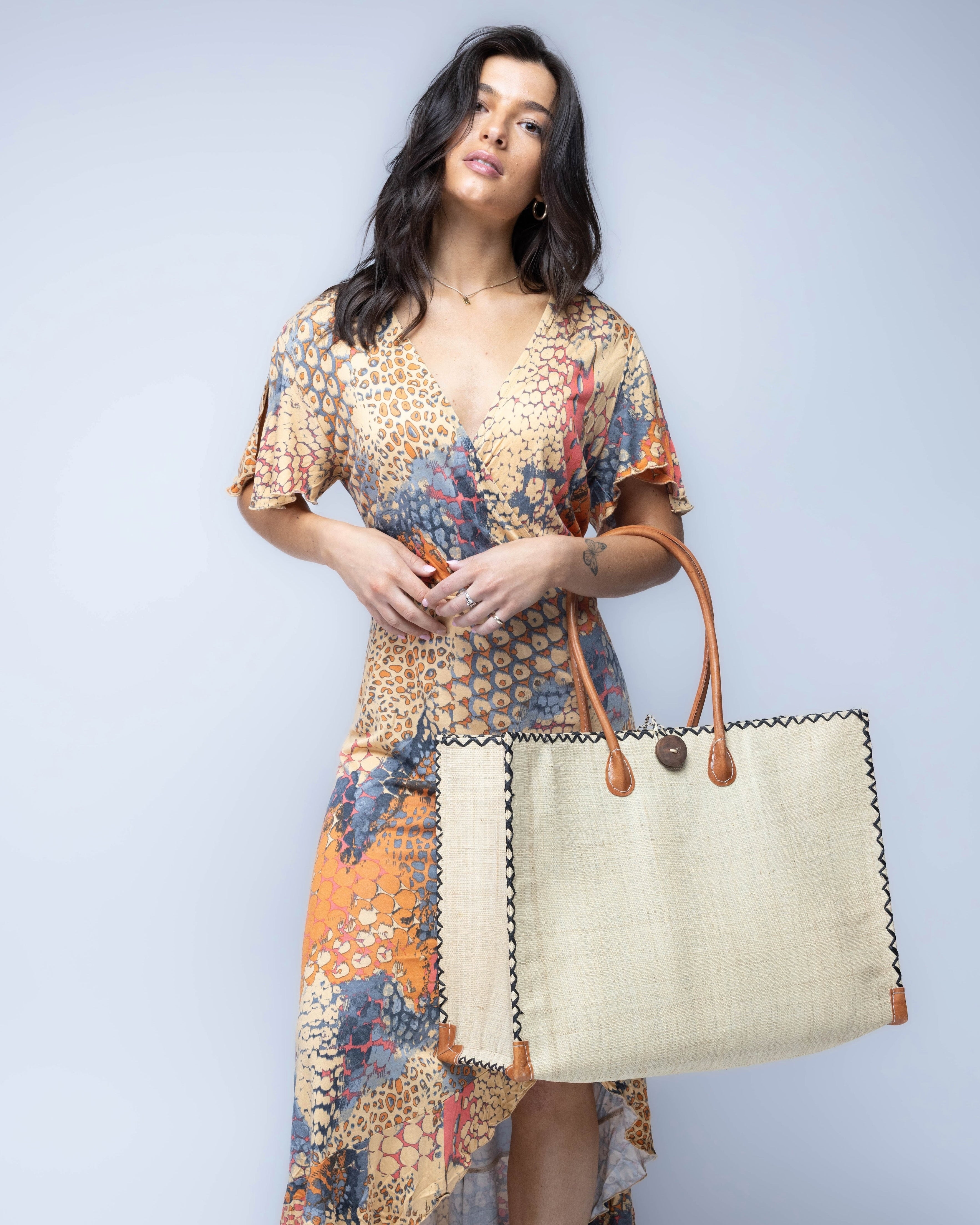 Model wearing Large Zafran Straw Beach Bag with Plastic Liner handmade loomed raffia in natural straw color with contrasting cross stitch edge binding, wood button closure, and leather handles & feet plus assorted print plastic lining - Shebobo