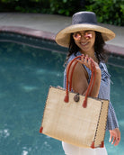 Model wearing Zafran Straw Beach Bag with Plastic Liner handmade loomed raffia in natural straw color with contrasting cross stitch edge binding, wood button closure, and leather handles & feet plus assorted print plastic lining - Shebobo (with 5" & 7" Wide Brim Two Tone Packable Straw Sun Hats)