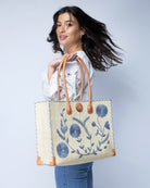Model wearing Zafran Natural & Denim Embroidered Straw Beach Bag with Floral Pattern handmade natural raffia palm fiber in a solid hue of natural straw color with asymmetrical floral leaf, vine, and flower pattern in denim retro/light/blue with matching cross-stitch edging, leather handles, and wooden button closure shoulder bag handbag tote - Shebobo
