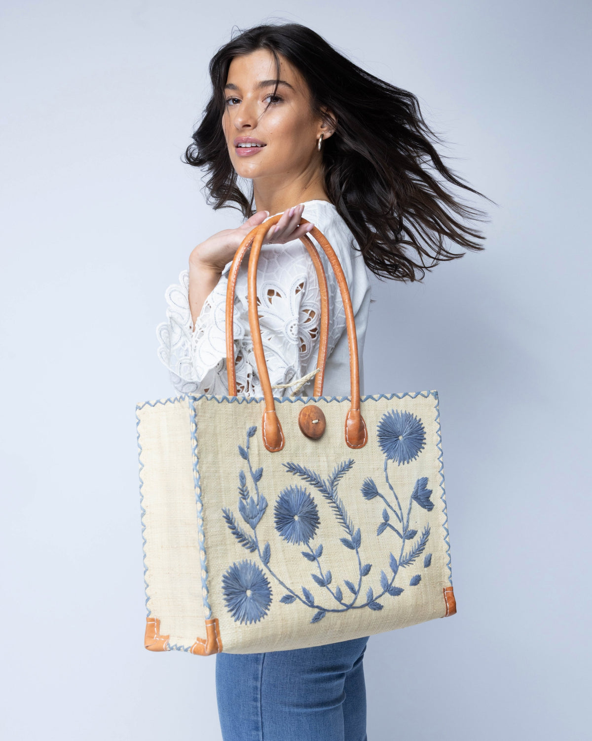 Model wearing Zafran Natural & Denim Embroidered Straw Beach Bag with Floral Pattern handmade natural raffia palm fiber in a solid hue of natural straw color with asymmetrical floral leaf, vine, and flower pattern in denim retro/light/blue with matching cross-stitch edging, leather handles, and wooden button closure shoulder bag handbag tote - Shebobo