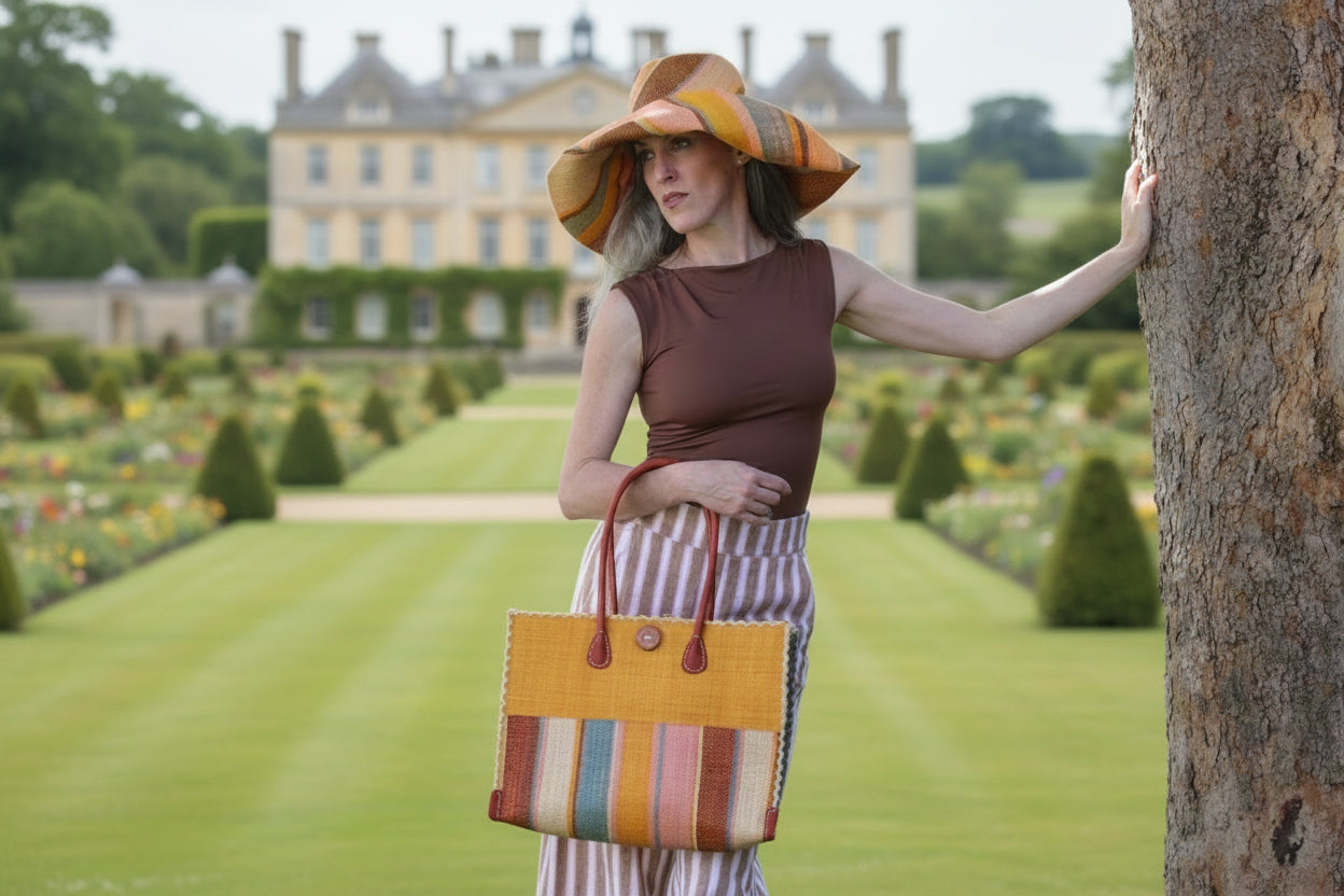 Model wearing Zafran Two Tone Wide Stripe Pink Blue Swirl Straw Beach Bag handmade loomed raffia palm fiber in a solid hue of Saffron yellow/orange on the top half of the bag and a multicolor multi-width vertical stripe pattern of black, soft/light/baby pink, light blue, saffron, and natural straw color on the bottom half of the bag with leather handles and feet and matching crosshatch stitch edging shoulder bag - Shebobo  (with Monica 5" & 7" Wide Brim Multicolor Stripe Pattern Packable Straw Sun Hats)
