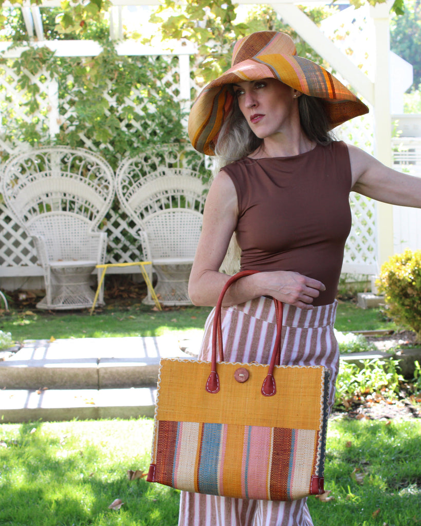 Model wearing Zafran Two Tone Wide Stripe Pink Blue Swirl Straw Beach Bag handmade loomed raffia palm fiber in a solid hue of Saffron yellow/orange on the top half of the bag and a multicolor multi-width vertical stripe pattern of black, soft/light/baby pink, light blue, saffron, and natural straw color on the bottom half of the bag with leather handles and feet and matching crosshatch stitch edging shoulder bag - Shebobo  (with Monica 5" & 7" Wide Brim Multicolor Stripe Pattern Packable Straw Sun Hats)