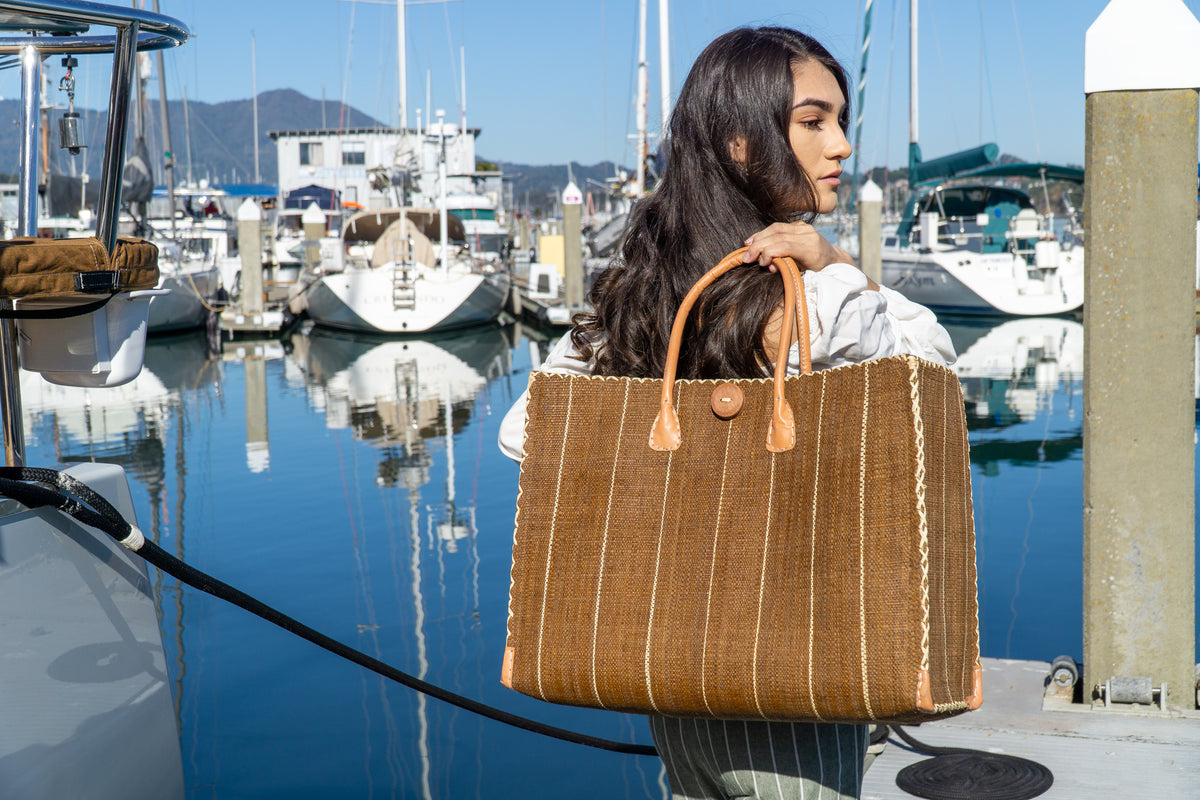 Model wearing Zafran Tobacco Pinstripes Large Straw Beach Bag with Plastic Liner handmade loomed raffia tote in wide bands of tobacco dark/cinnamon/brown with narrow bands of natural straw color that make a vertical pinstripe pattern with assorted print plastic lining and leather handles xl bag - Shebobo