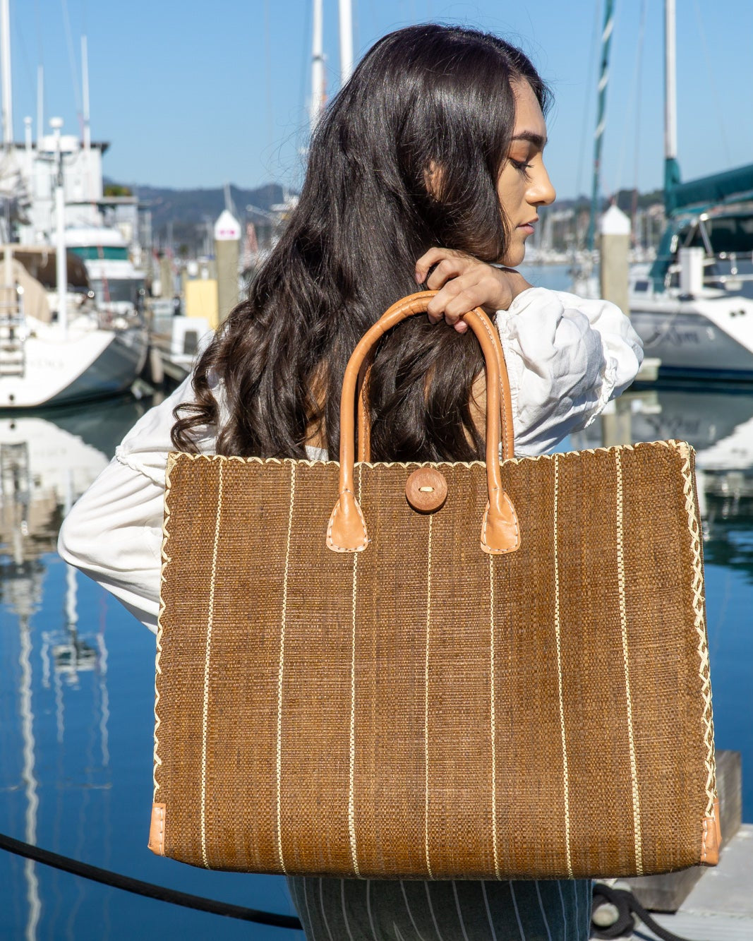 Model wearing Zafran Tobacco Pinstripes Large Straw Beach Bag with Plastic Liner handmade loomed raffia tote in wide bands of tobacco dark/cinnamon/brown with narrow bands of natural straw color that make a vertical pinstripe pattern with assorted print plastic lining and leather handles xl bag - Shebobo
