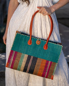 Model wearing Zafran Two Tone Wide Stripe Revel Swirl Straw Beach Bag handmade loomed raffia palm fiber in a solid hue of Teal dark/blue/green on the top half of the bag and a multicolor multi-width vertical stripe pattern of black, cinnamon tobacco/dark/brown, teal, coral orange/red, fuchsia hot/bright/barbie/pink, and natural straw color on the bottom half of the bag with leather handles and feet and matching crosshatch stitch edging shoulder bag - Shebobo