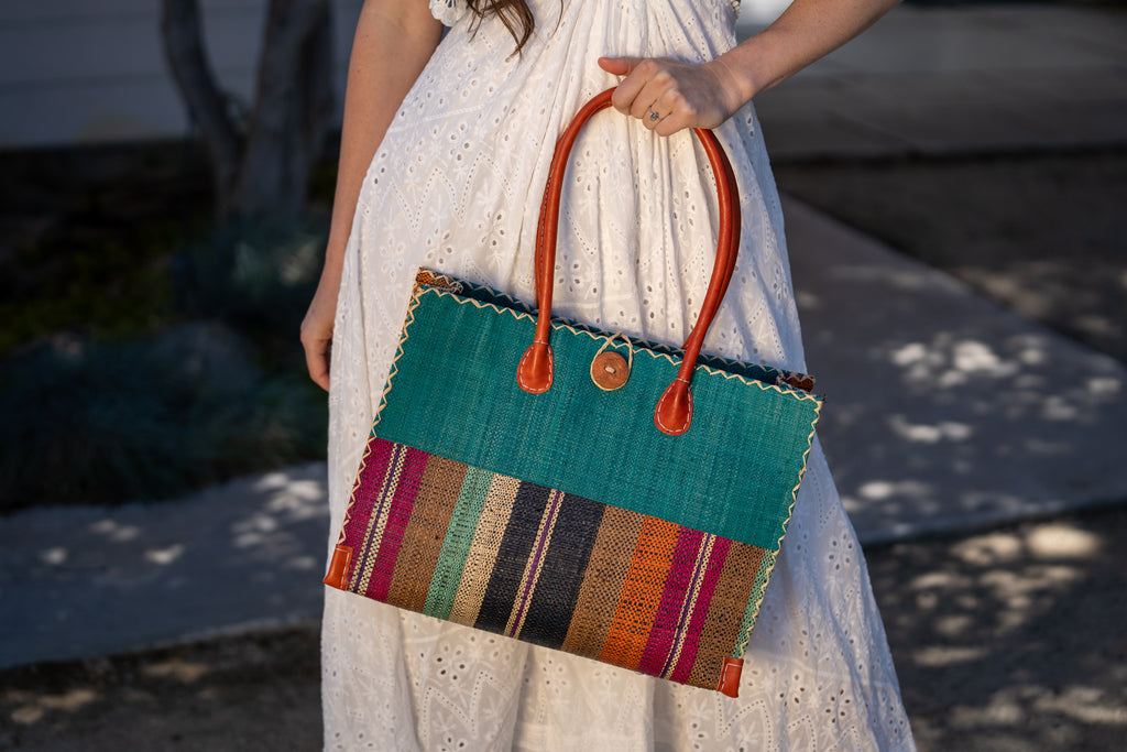 Model wearing Zafran Two Tone Wide Stripe Revel Swirl Straw Beach Bag handmade loomed raffia palm fiber in a solid hue of Teal dark/blue/green on the top half of the bag and a multicolor multi-width vertical stripe pattern of black, cinnamon tobacco/dark/brown, teal, coral orange/red, fuchsia hot/bright/barbie/pink, and natural straw color on the bottom half of the bag with leather handles and feet and matching crosshatch stitch edging shoulder bag - Shebobo