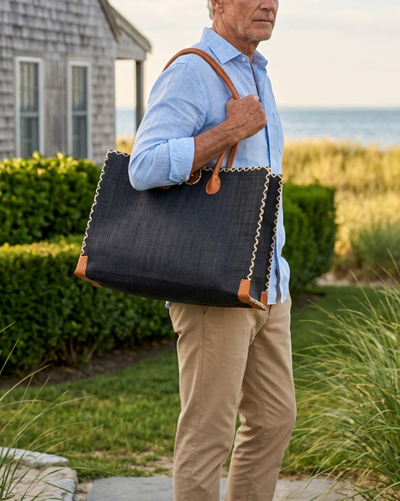 Model wearing Zafran Straw Beach Bag with Plastic Liner handmade loomed raffia in a solid hue of black with contrasting natural straw color cross stitch edge binding, wood button closure, and leather handles & feet plus assorted print plastic lining - Shebobo
