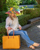 Model wearing Zafran Straw Beach Bag with Plastic Liner handmade loomed raffia in a solid hue of saffron yellow/orange with contrasting natural straw color cross stitch edge binding, wood button closure, and leather handles & feet plus assorted print plastic lining - Shebobo (with 5" & 7" Wide Brim Carmalita Multicolor Stripe Pattern Packable Straw Sun Hats and Lizzie Straw Sandal)