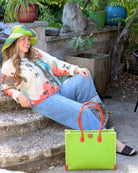 Model wearing Zafran Straw Beach Bag with Plastic Liner handmade loomed raffia in a solid hue of Midori lime/bright/neon/green with contrasting natural straw color cross stitch edge binding, wood button closure, and leather handles & feet plus assorted print plastic lining - Shebobo (with Monica 5" & 7" Wide Brim Multicolor Stripe Pattern Packable Straw Sun Hats)
