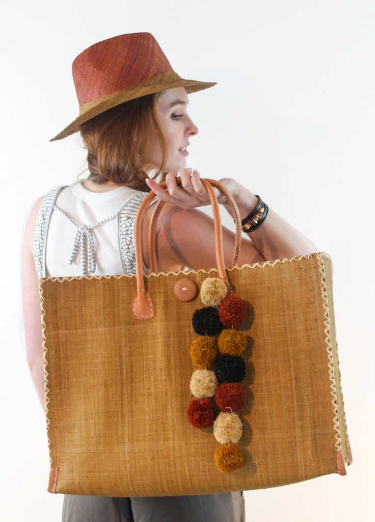 Model wearing Model wearing Zafran Large Straw Beach Bag with Plastic Liner handmade loomed raffia in cinnamon/tobacco/brown color with contrasting cross stitch edge binding, wood button closure, and leather handles & feet plus assorted print plastic lining with Panama Two Tone Unisex Straw Hat in Cinnamon/Caramel plus Strand Pompom Charm Embellishment in Boho Browns Multicolor Multi Raffia Poufs - Shebobo