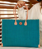 Model wearing Zafran Straw Beach Bag with Plastic Liner handmade loomed raffia in a solid hue of Turquoise blue/green with contrasting natural straw color cross stitch edge binding, wood button closure, and leather handles & feet plus assorted print plastic lining - Shebobo