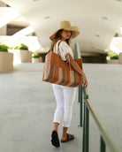 Model wearing Zafran Two Tone Wide Stripe Blush Swirl Straw Beach Bag handmade loomed raffia palm fiber in a solid hue of blush orange/pink on the top half of the bag and a multicolor multi-width vertical stripe pattern of blush, grey, and natural straw color on the bottom half of the bag with leather handles and feet and matching crosshatch stitch edging shoulder bag - Shebobo (with 7" Wide Brim Natural Packable Straw Sun Hat)