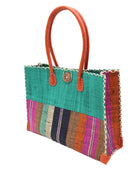 Side view Zafran Two Tone Wide Stripe Revel Swirl Straw Beach Bag handmade loomed raffia palm fiber in a solid hue of Teal dark/blue/green on the top half of the bag and a multicolor multi-width vertical stripe pattern of black, cinnamon tobacco/dark/brown, teal, coral orange/red, fuchsia hot/bright/barbie/pink, and natural straw color on the bottom half of the bag with leather handles and feet and matching crosshatch stitch edging shoulder bag - Shebobo