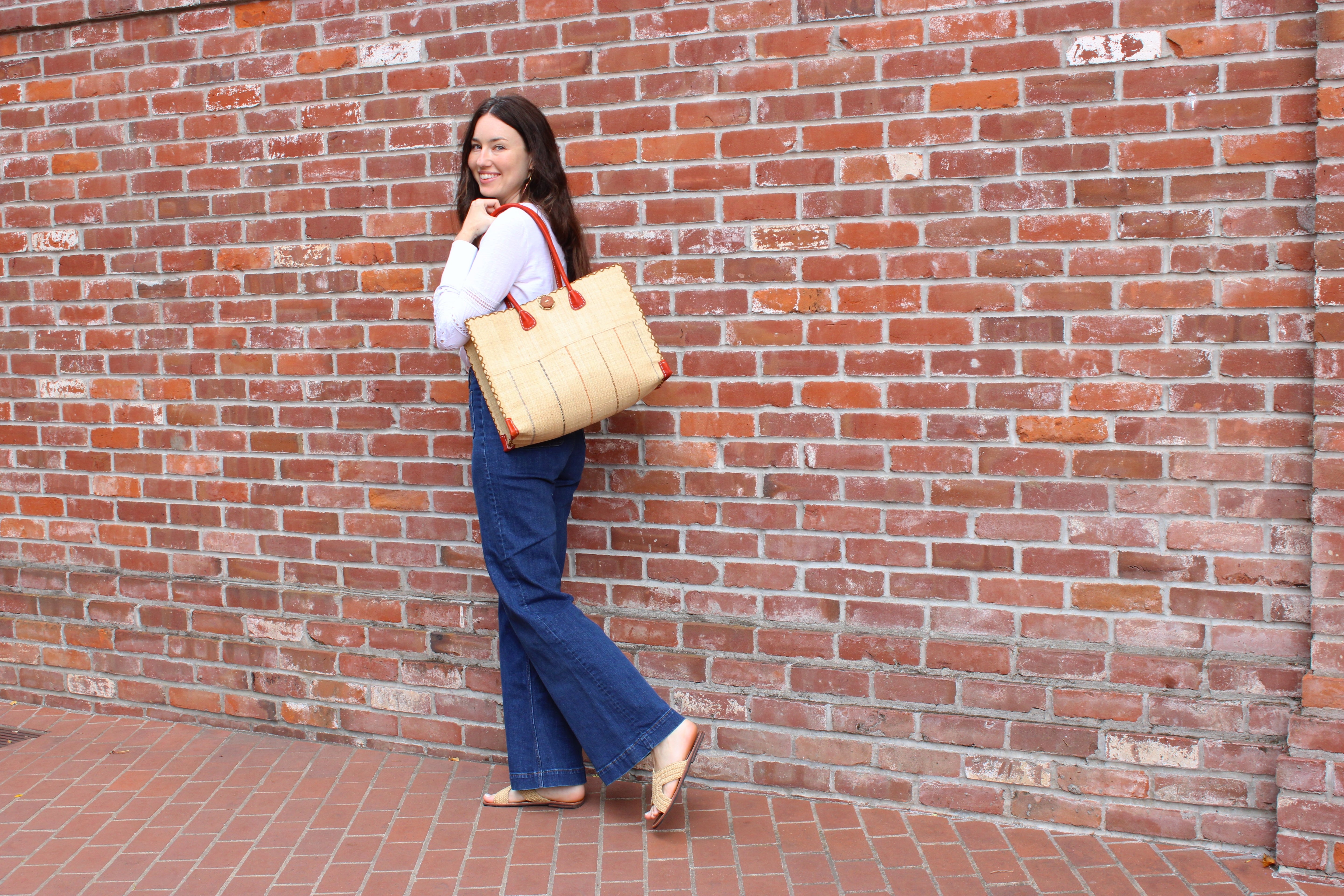 Model wearing Zafran Two Tone Wide Stripe Neutrals Pinstripe Straw Beach Bag handmade loomed raffia palm fiber in a solid hue of natural straw color on the top half of the bag and a multicolor multi-width vertical pinstripe stripe pattern with thin bands of black, blush pink/orange, cinnamon tobacco/dark/brown, and grey with wide bands of natural straw color on the bottom half of the bag with leather handles and feet and matching crosshatch stitch edging shoulder bag - Shebobo