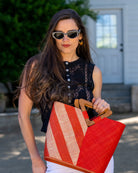 Model wearing Zuki Two Tone Coral Straw Handbag With Wood Triangle Handle handmade loomed raffia palm fiber in a two tone pattern with the left half of the bag even width stripes of coral orange/red and natural straw color oriented as a diagonal stripe and the right half of the bag a solid hue of coral with carved wood handles - Shebobo
