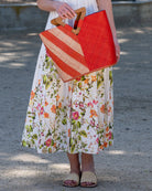 Model wearing Zuki Two Tone Coral Straw Handbag With Wood Triangle Handle handmade loomed raffia palm fiber in a two tone pattern with the left half of the bag even width stripes of coral orange/red and natural straw color oriented as a diagonal stripe and the right half of the bag a solid hue of coral with carved wood handles - Shebobo (with Lulu Macrame Sandal)