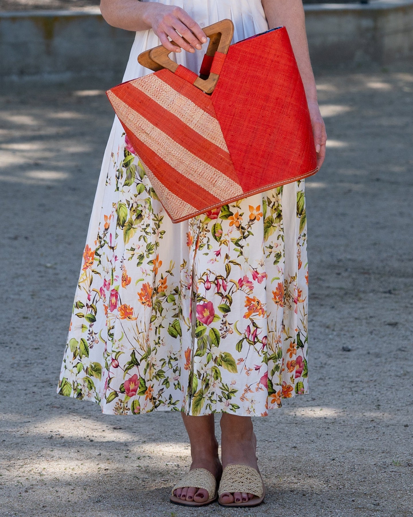 Model wearing Zuki Two Tone Coral Straw Handbag With Wood Triangle Handle handmade loomed raffia palm fiber in a two tone pattern with the left half of the bag even width stripes of coral orange/red and natural straw color oriented as a diagonal stripe and the right half of the bag a solid hue of coral with carved wood handles - Shebobo (with Lulu Macrame Sandal)