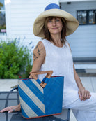 Model wearing Zuki Two Tone Denim Straw Handbag With Wood Triangle Handle handmade loomed raffia palm fiber in a two tone pattern with the left half of the bag even width stripes of denim retro/light/blue and natural straw color oriented as a diagonal stripe and the right half of the bag a solid hue of denim with carved wood handles - Shebobo (Monica 5" & 7" Wide Brim Two Tone Packable Straw Sun Hats)