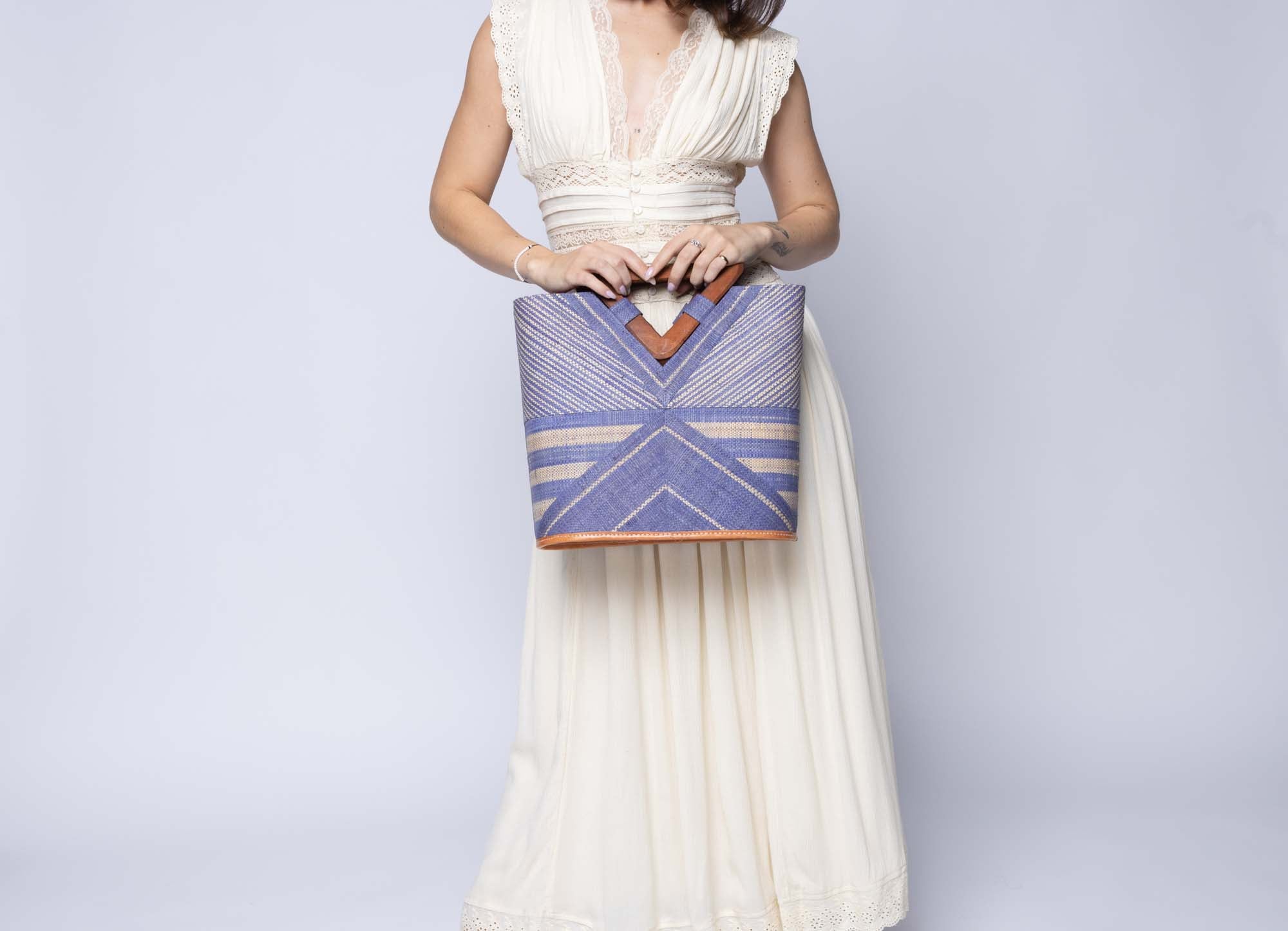 Model wearing Mariposa Zuki Raffia Straw Bucket Handbag with Wood Triangle Handles  handmade loomed raffia palm fiber in a multi paneled pattern design of pinstripe, melange, and wide stripe combinations geometrically radiating from the center point of the bag outwards in a two tone color combination of Denim and Natural straw color with a solid hue of color on the back of the bag plus carved wooden handles purse - Shebobo