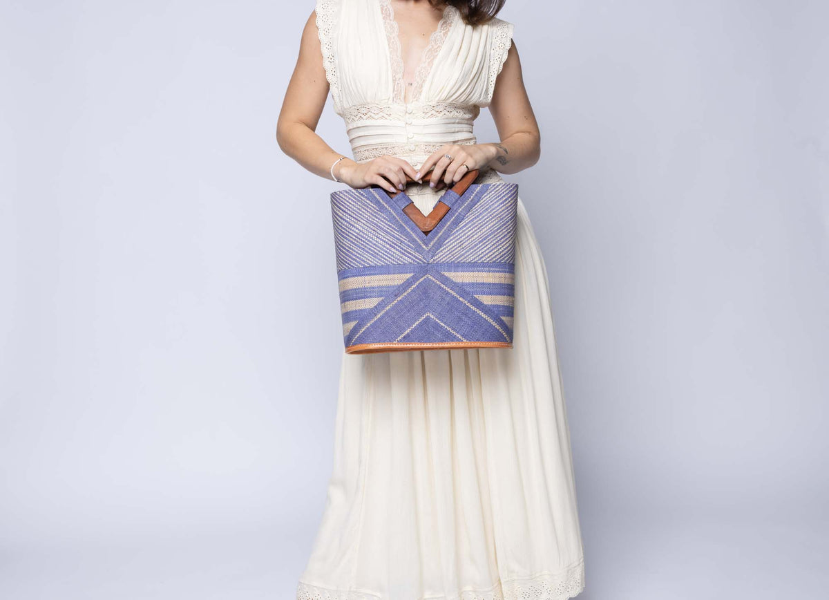 Model wearing Mariposa Zuki Raffia Straw Bucket Handbag with Wood Triangle Handles  handmade loomed raffia palm fiber in a multi paneled pattern design of pinstripe, melange, and wide stripe combinations geometrically radiating from the center point of the bag outwards in a two tone color combination of Denim and Natural straw color with a solid hue of color on the back of the bag plus carved wooden handles purse - Shebobo
