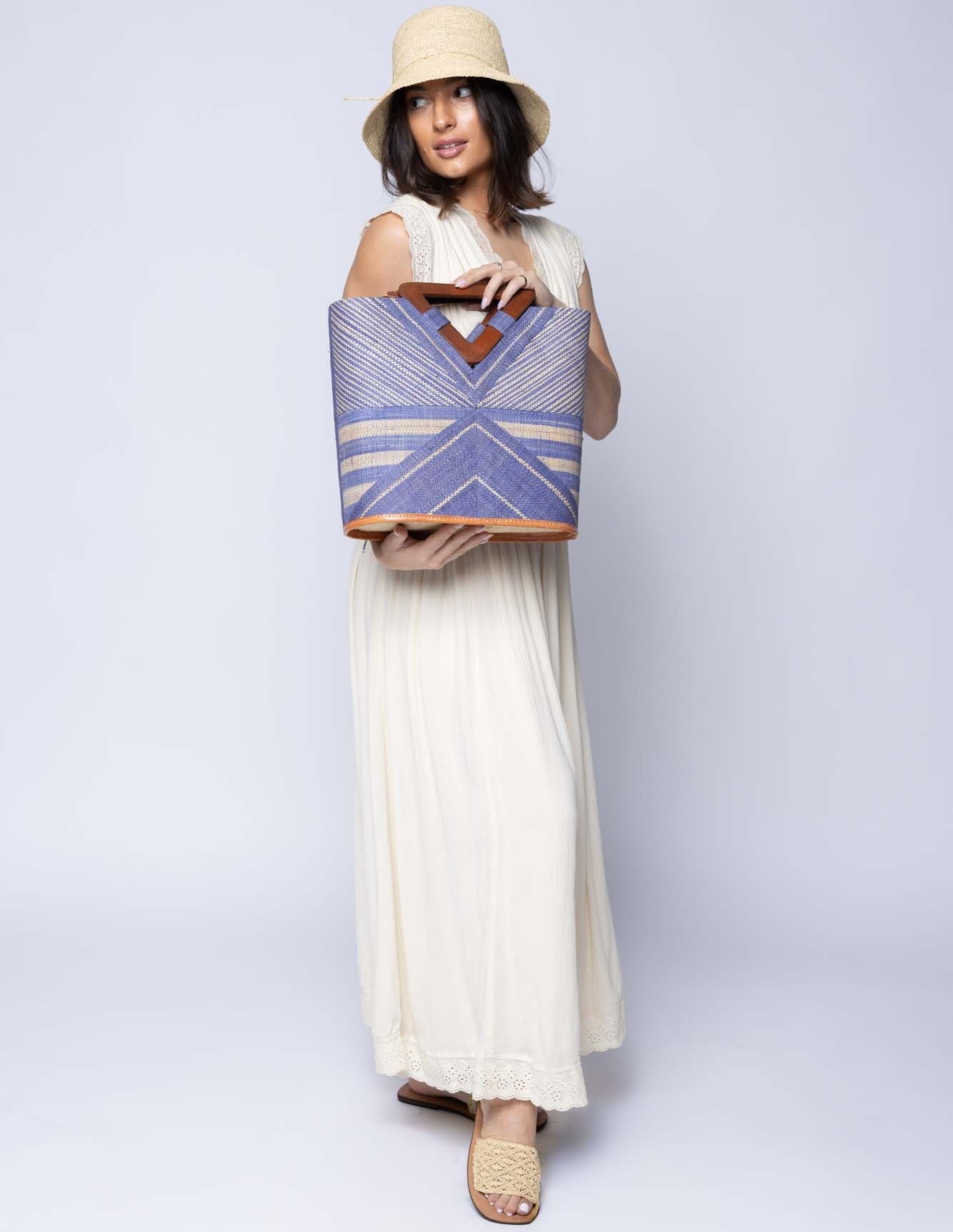 Model wearing Mariposa Zuki Raffia Straw Bucket Handbag with Wood Triangle Handles  handmade loomed raffia palm fiber in a multi paneled pattern design of pinstripe, melange, and wide stripe combinations geometrically radiating from the center point of the bag outwards in a two tone color combination of Denim and Natural straw color with a solid hue of color on the back of the bag plus carved wooden handles purse - Shebobo