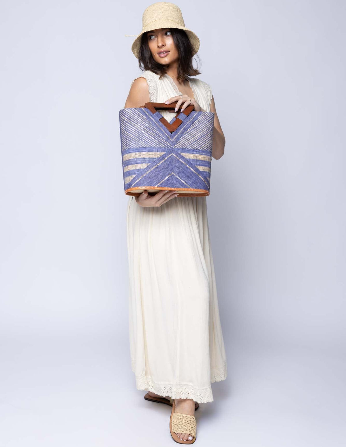 Model wearing Mariposa Zuki Raffia Straw Bucket Handbag with Wood Triangle Handles  handmade loomed raffia palm fiber in a multi paneled pattern design of pinstripe, melange, and wide stripe combinations geometrically radiating from the center point of the bag outwards in a two tone color combination of Denim and Natural straw color with a solid hue of color on the back of the bag plus carved wooden handles purse - Shebobo