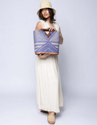 Model wearing Mariposa Zuki Raffia Straw Bucket Handbag with Wood Triangle Handles  handmade loomed raffia palm fiber in a multi paneled pattern design of pinstripe, melange, and wide stripe combinations geometrically radiating from the center point of the bag outwards in a two tone color combination of Denim and Natural straw color with a solid hue of color on the back of the bag plus carved wooden handles purse - Shebobo