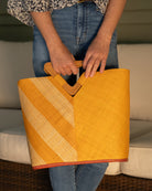 Model wearing Zuki Two Tone Saffron Straw Handbag With Wood Triangle Handle handmade loomed raffia palm fiber in a two tone pattern with the left half of the bag even width stripes of saffron yellow/orange and natural straw color oriented as a diagonal stripe and the right half of the bag a solid hue of saffron with carved wood handles - Shebobo