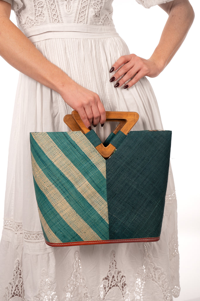 Model wearing Zuki Two Tone Teal Straw Handbag With Wood Triangle Handle handmade loomed raffia palm fiber in a two tone pattern with the left half of the bag even width stripes of teal dark/blue/green and natural straw color oriented as a diagonal stripe and the right half of the bag a solid hue of teal with carved wood handles - Shebobo