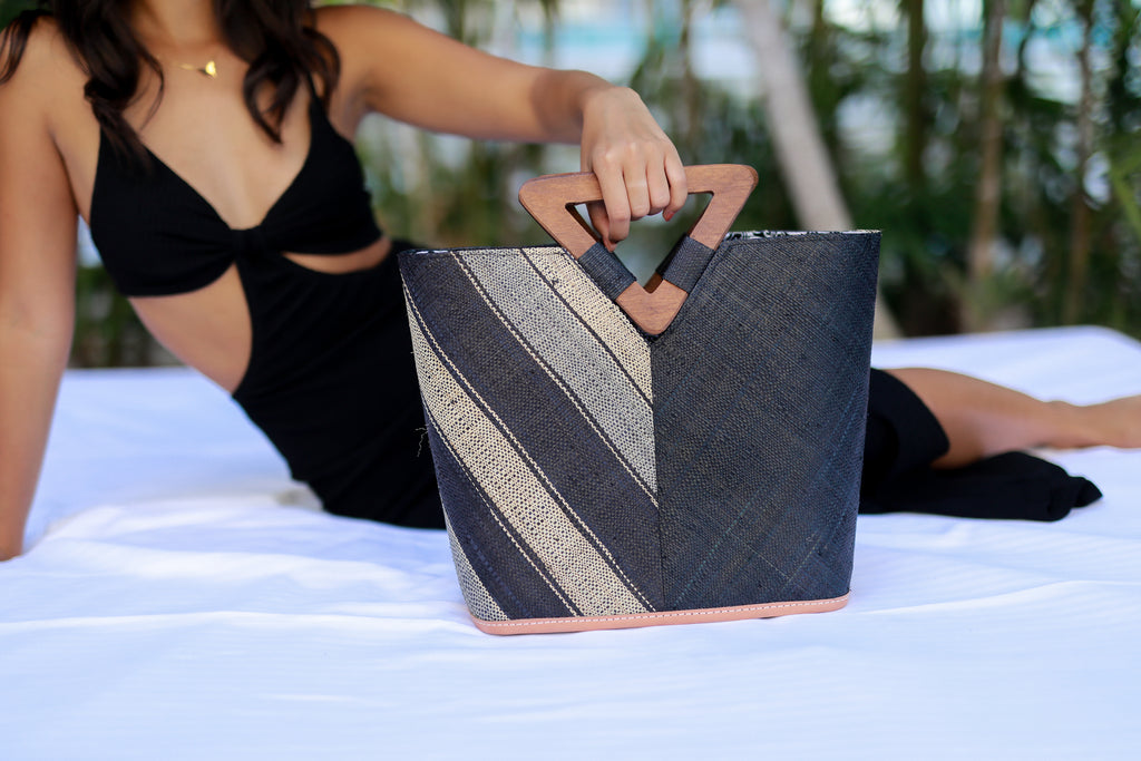 Model wearing Zuki Two Tone Black Swirl Straw Handbag With Wood Triangle Handle handmade loomed raffia vertically halved visually with one side of multicolor black, grey, and natural multi width stripe pattern on a diagonal with the other half solid black boho chic purse bag - Shebobo