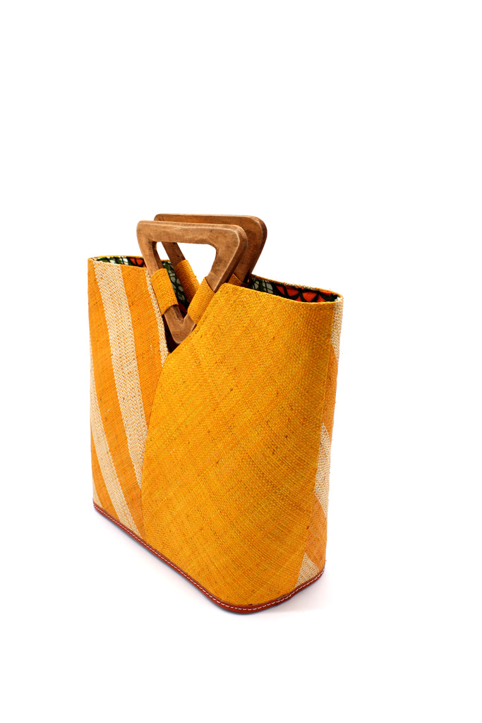 Side View Zuki Two Tone Saffron Straw Handbag With Wood Triangle Handle handmade loomed raffia palm fiber in a two tone pattern with the left half of the bag even width stripes of saffron yellow/orange and natural straw color oriented as a diagonal stripe and the right half of the bag a solid hue of saffron with carved wood handles - Shebobo