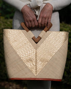 Model wearing Zuki Wynwood Natural Straw Handbag with Metallic Detailing & Wood Triangle Handle handmade loomed raffia palm fiber in a solid hue of natural straw color with incorporated vegan leather  gold metallic strands in a V pattern across the front and back of the bag with assorted print African wax cloth liners and carved wooden triangle handle purse - Shebobo