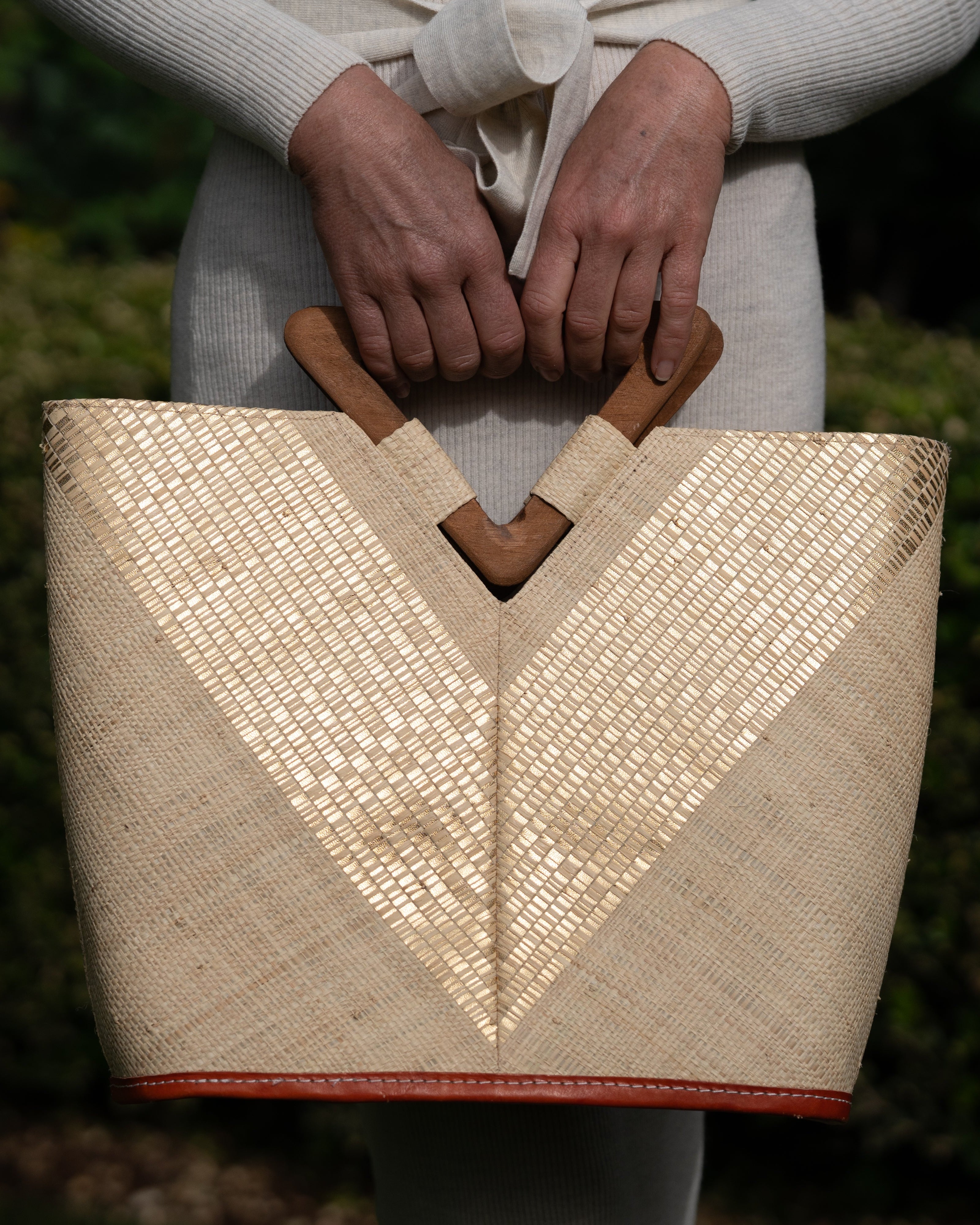 Model wearing Zuki Wynwood Natural Straw Handbag with Metallic Detailing & Wood Triangle Handle handmade loomed raffia palm fiber in a solid hue of natural straw color with incorporated vegan leather  gold metallic strands in a V pattern across the front and back of the bag with assorted print African wax cloth liners and carved wooden triangle handle purse - Shebobo