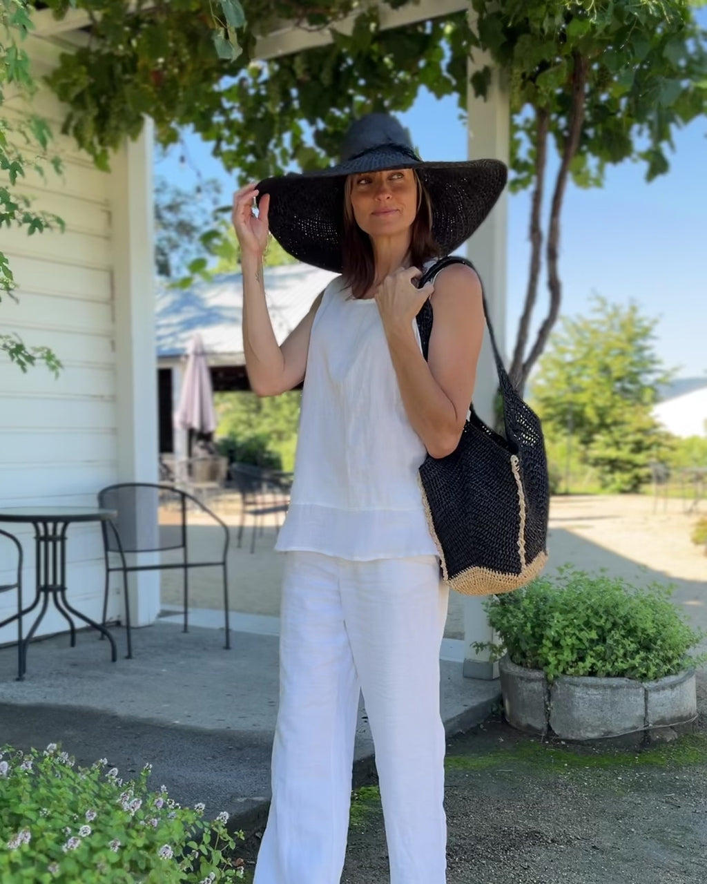 Model wearing Sea See Black Straw Shoulder Bag handmade woven raffia palm fiber in a solid hue of black making a diagonal link pattern with  natural straw color crochet circular bottom and vertical edging along the side panels creating a two tone multicolor handbag/beach bag/purse - Shebobo (with Natasha 7" Wide Brim Woven XL Straw Sun Hats)