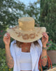 Model wearing 5.5" Brim Gigi Natural and Cinnamon Floral Embroidered Crochet Straw Sun Hats handmade crochet natural raffia palm fiber in a solid hue of natural straw color making a concentric pattern from the center top of the flattened crown spiraling out to the edge of the brim with matching adjustable raffia braid hat band and cinnamon dark brown floral pattern with leaves embroidered on the brim - Shebobo