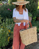 Model wearing Flower Power Natural Crochet Straw Basket Bag handmade from natural raffia palm fiber in a granny square-esque geometric floral pattern with flower detailing projecting from main crochet textile in a natural straw color with leather handles handbag purse - Shebobo (with Gigi 5.5" Brim Floral Embroidered Crochet Straw Sun Hats)