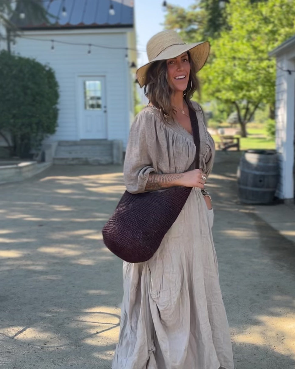 Model wearing Model wearing Kiki Crochet Straw Crossbody Bag handmade natural raffia palm fiber in a crochet pattern of subtle linked bands in a solid hue of dark chocolate brown purse - Shebobo (with Rachel 4" Brim Crochet Straw Sun Hats with Colored Edge)