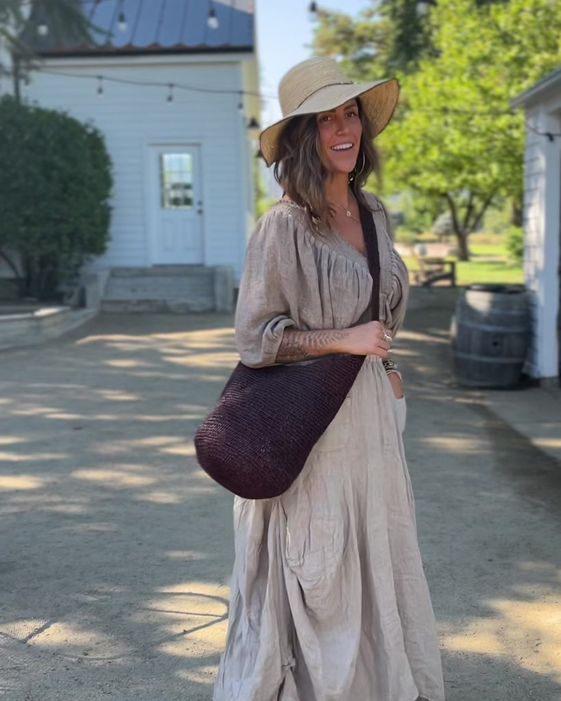 Model wearing Model wearing Kiki Crochet Straw Crossbody Bag handmade natural raffia palm fiber in a crochet pattern of subtle linked bands in a solid hue of dark chocolate brown purse - Shebobo (with Rachel 4" Brim Crochet Straw Sun Hats with Colored Edge)