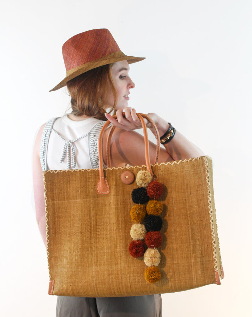 Model wearing beach bag adorned with Strand Pompoms Charm Embellishment multiple handmade multicolor caramel brown, black, natural, and saffron yellow raffia pompoms attached to raffia cord - great for beach bag flair or decor - Shebobo